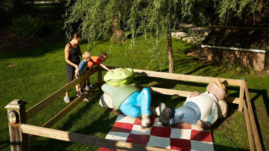 An adult and two children look at a sculpture of an oversized frog resting on a picnic blanket.